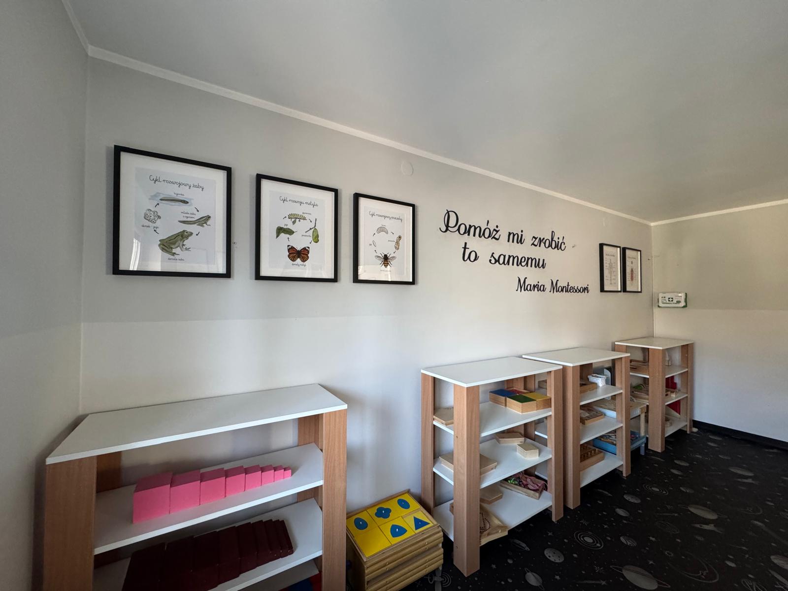 Sala, gdzie wzdłuż ścian ustawione są niskie, drewniane regały z białymi blatami, na których znajdują się różnorodne materiały i zabawki Montessori. Na ścianach wiszą oprawione plansze edukacyjne przedstawiające zwierzęta i rośliny oraz cytat Marii Montessori: "Pomóż mi zrobić to samemu".