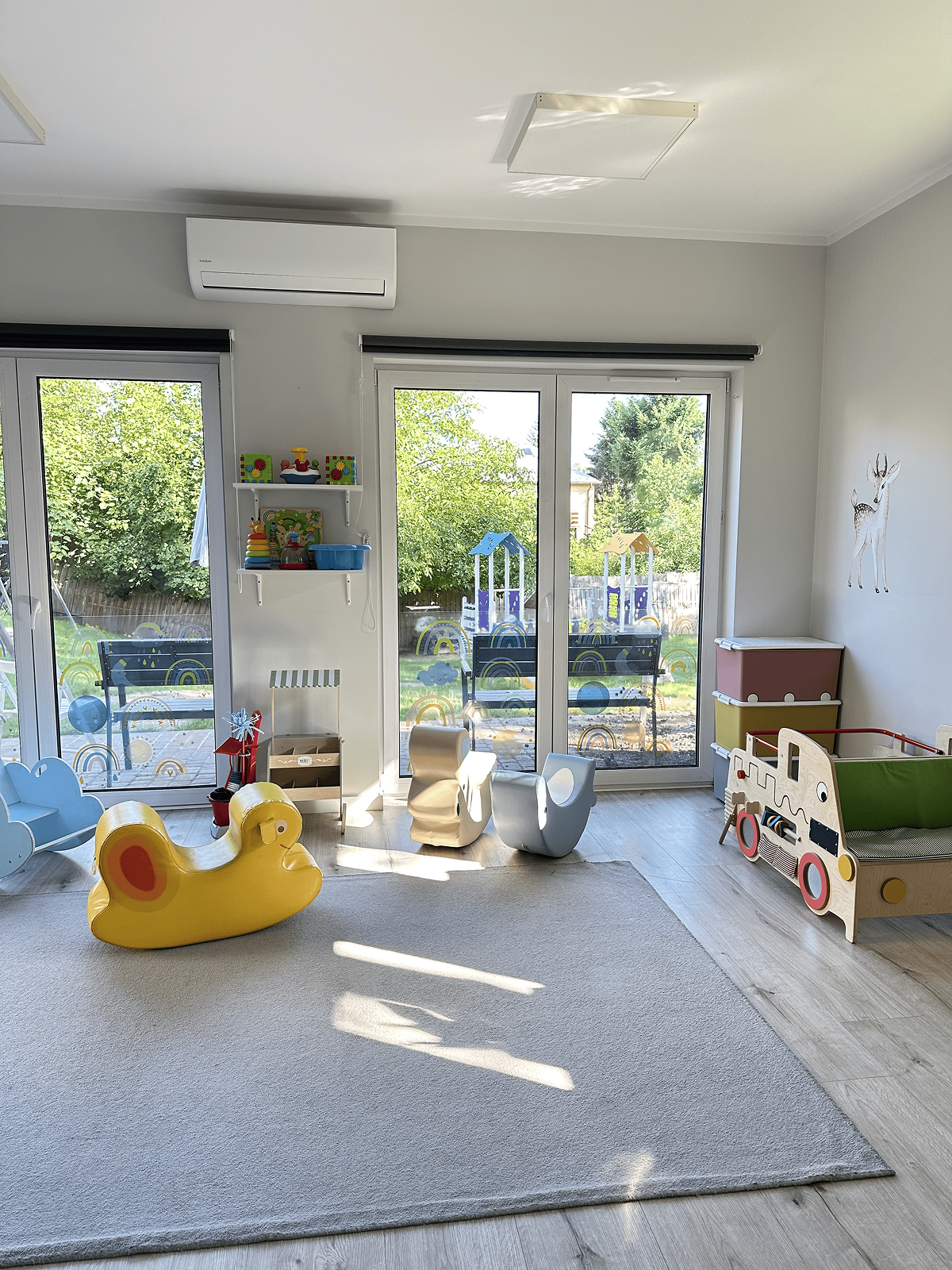 Przestronna sala zabaw. W sali znajdują się drewniane jeździki w kształcie zwierząt, drewniana sensoryczna ciężarówka w stylu Montessori, białe półki z pojemnikami i zabawkami. Na ścianach widoczne są naklejki z motywami zwierząt i lasu.