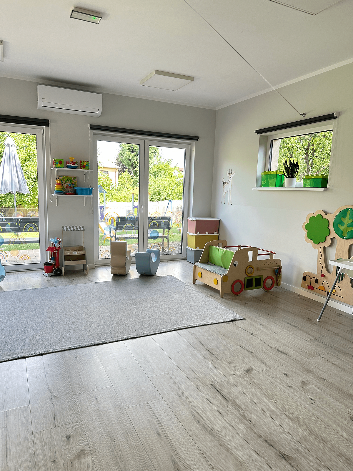 Sala zabaw z drewnianymi bujakami oraz drewnianymi akcesoriami Montessori- samochodem oraz drzewem na ścianie.